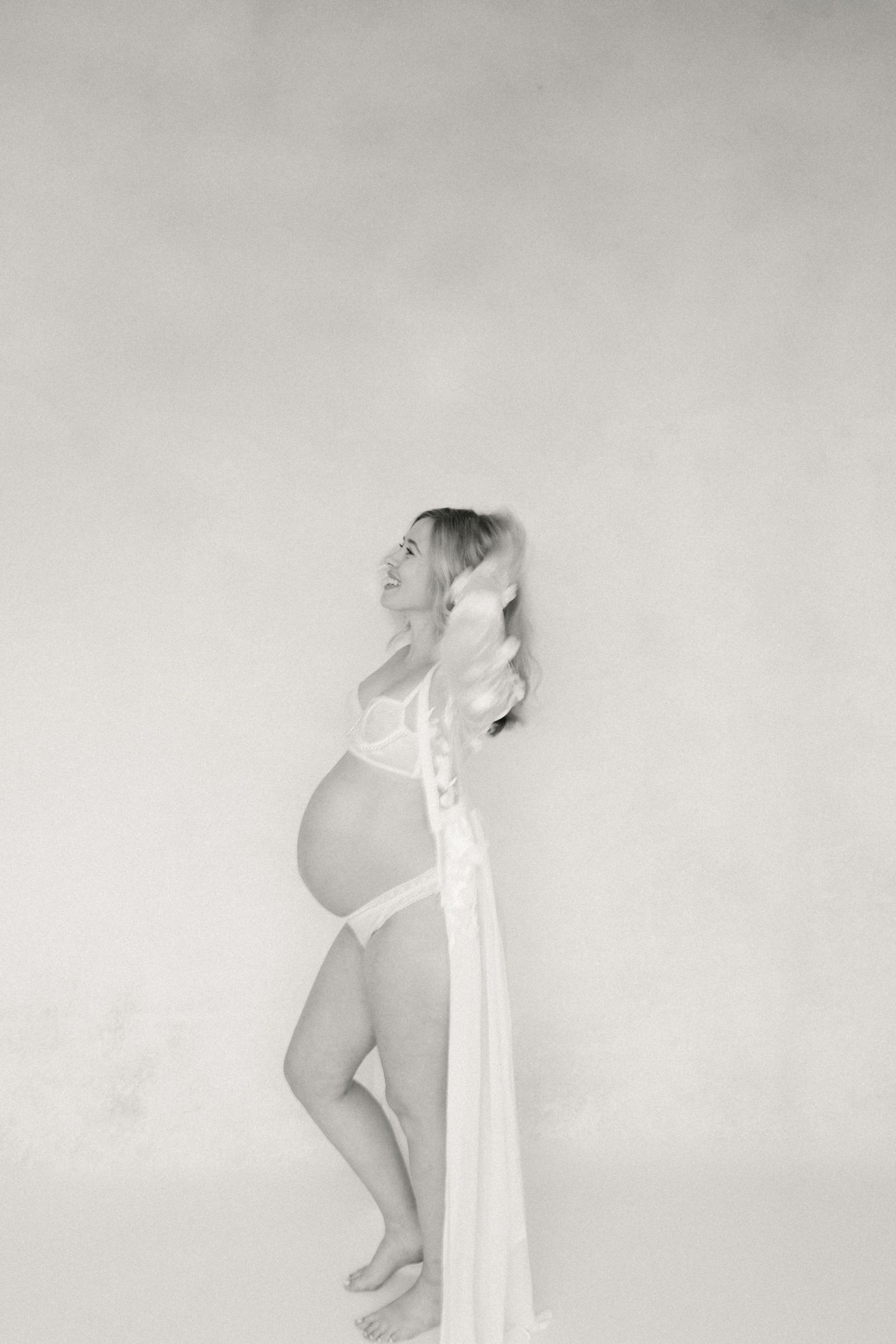 Black and white artistic maternity photo showing side profile of woman in flowing white fabric against plain background.