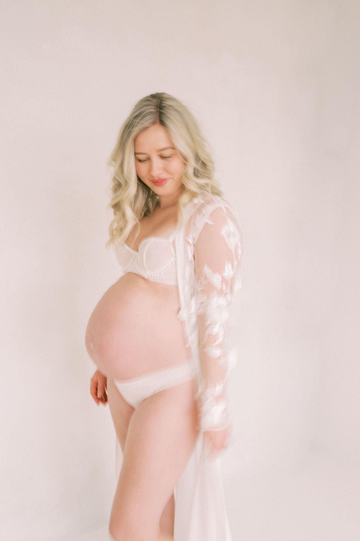 Ethereal maternity photo with blonde woman in white lace gown against soft pink background.