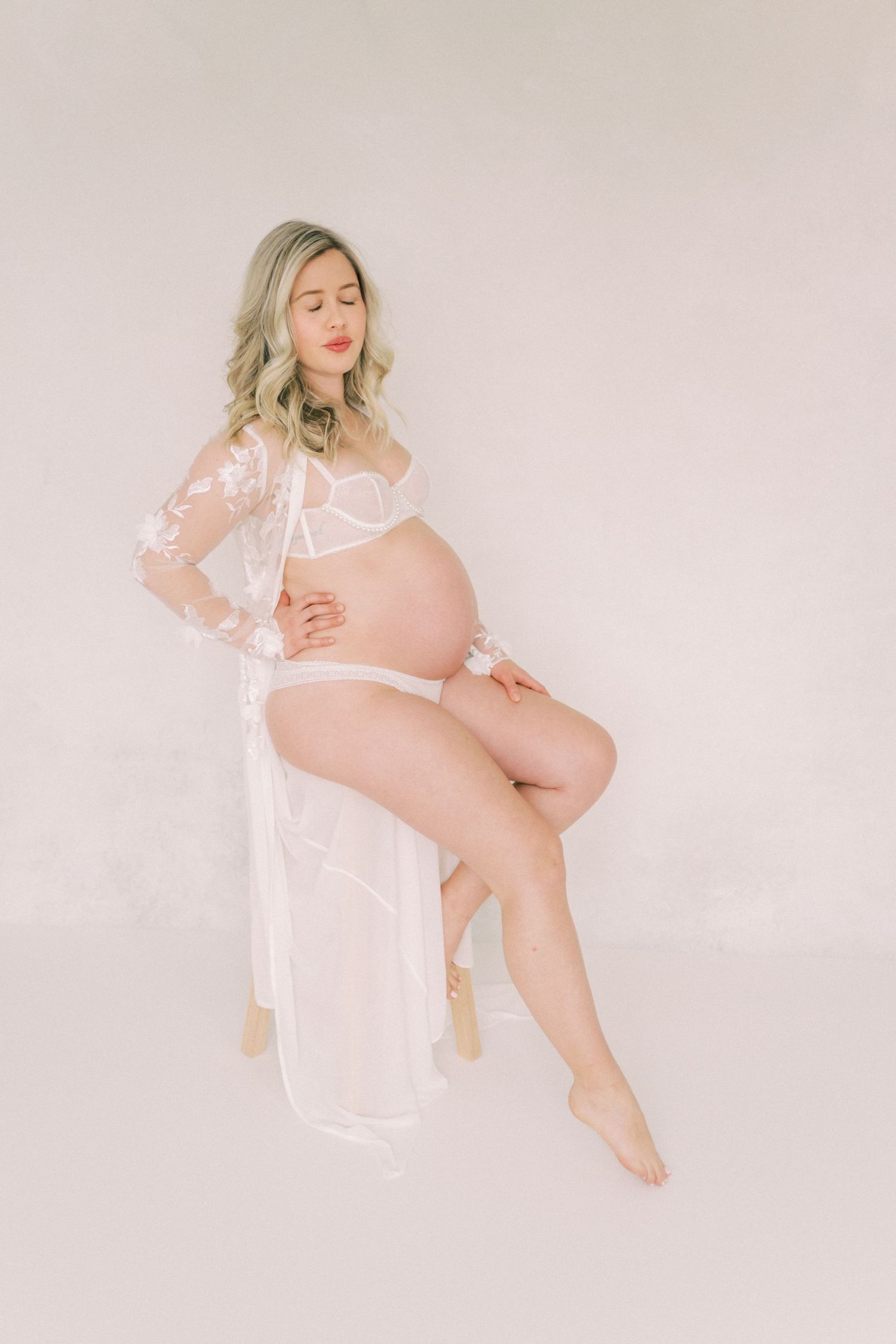 Maternity portrait with soft white lace robe and lingerie against neutral beige background.