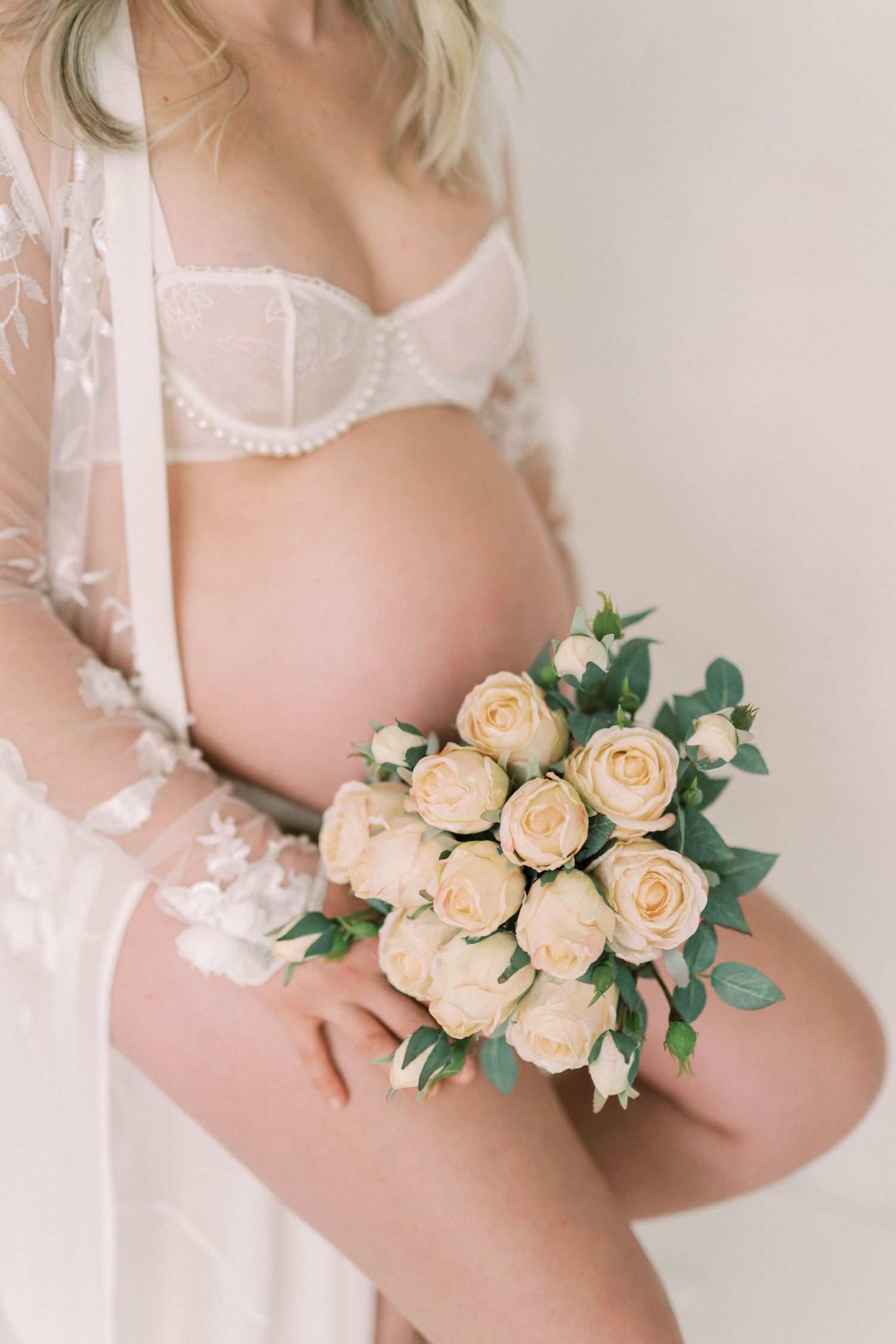 Beautiful maternity photo featuring cream roses bouquet and delicate white lace lingerie against soft neutral background.
