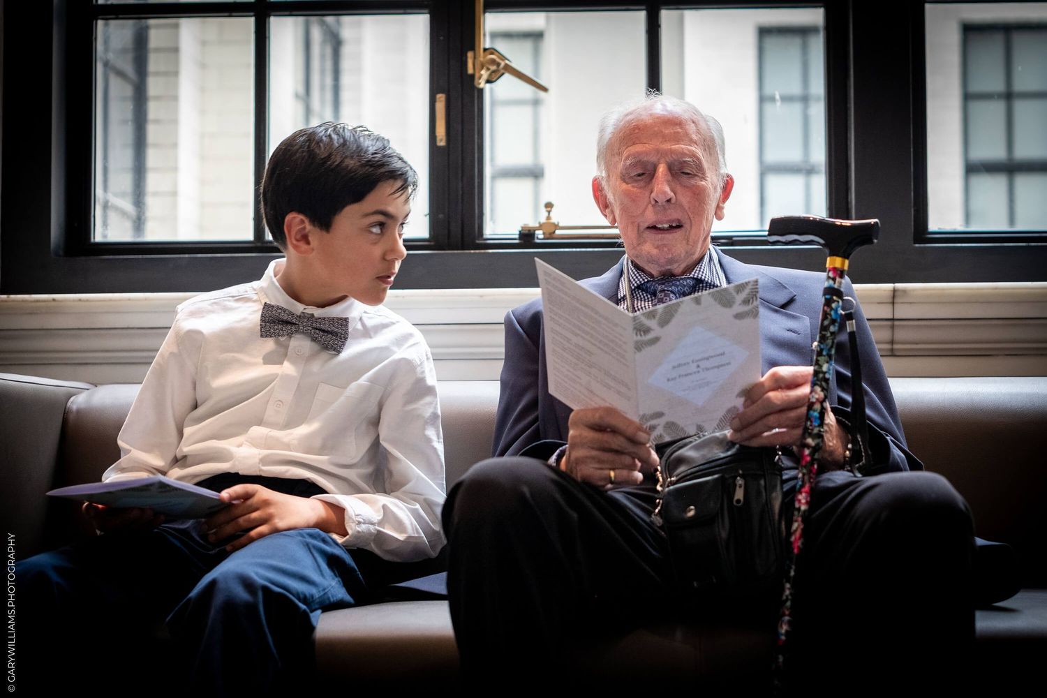 Two people sit together on a bench, with one reading papers while wearing a bowtie and the other looking on.