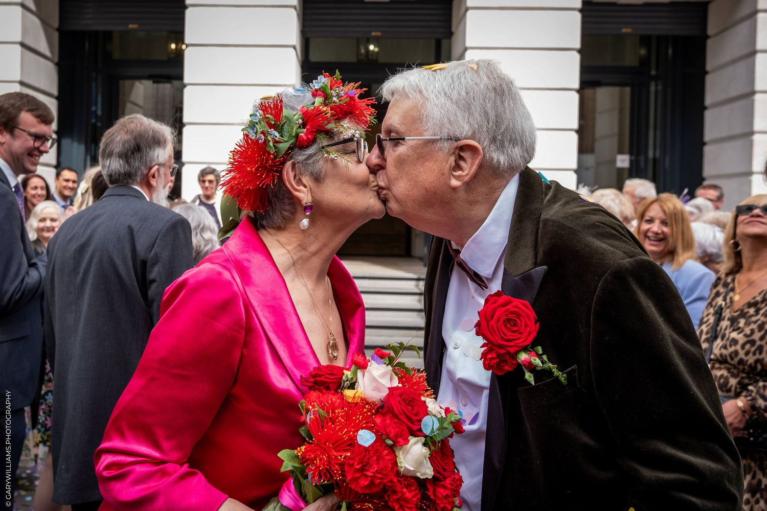 Senior couple wearing formal attire and red flowers share a kiss outside a building with onlookers.