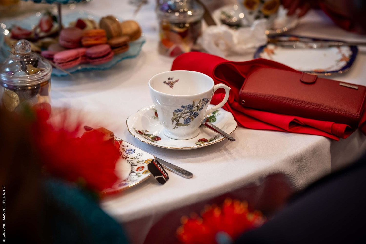 An elegant tea cup and saucer with floral design sits on a white tablecloth next to a red clutch purse.
