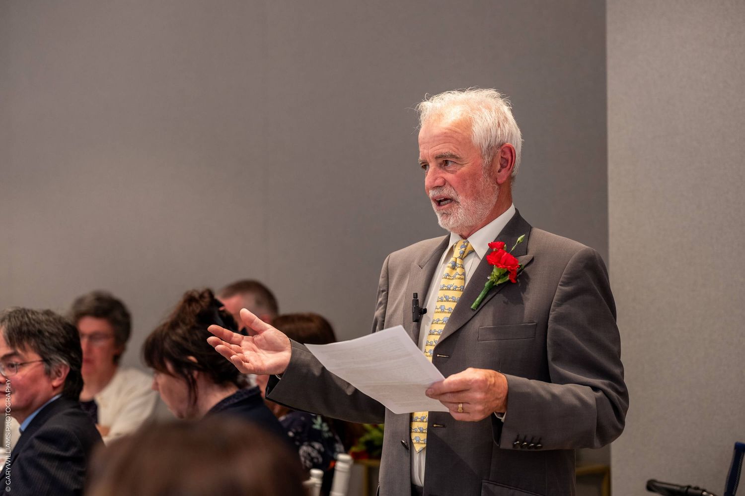 A well-dressed speaker in a gray suit with a red boutonniere holds papers while addressing an audience at an event.
