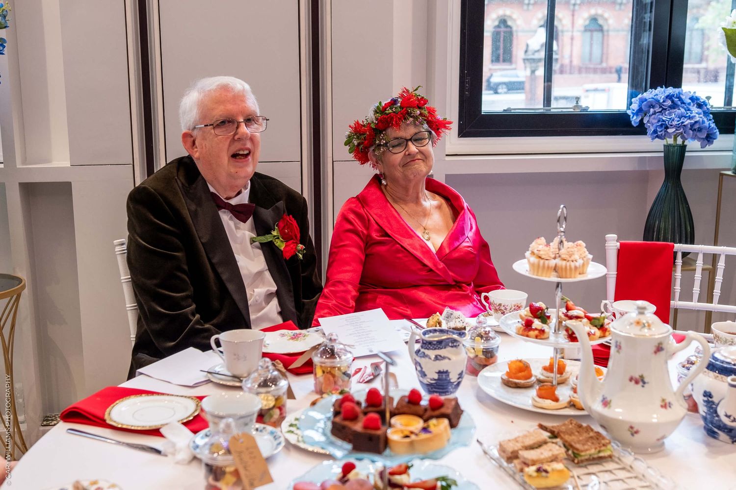 Two guests dressed in formal attire enjoying a festive afternoon tea with scones and treats at an elegant table setting.