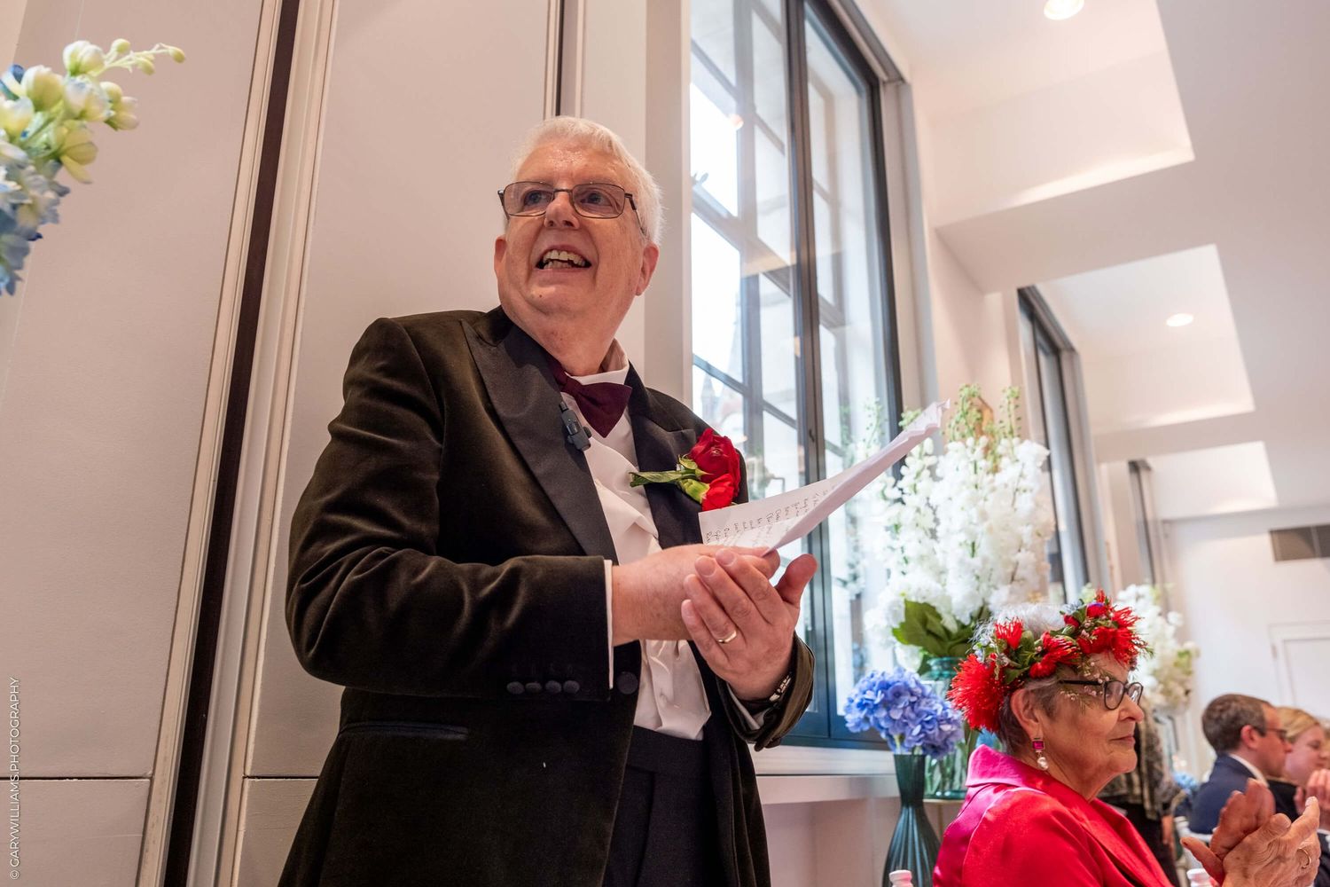 A person in a dark suit jacket with a red boutonniere smiles while holding papers during an indoor event.