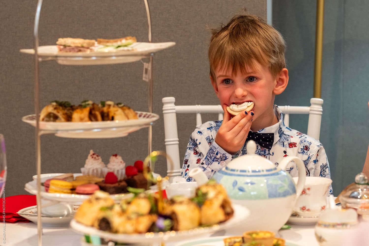 Young child sitting at table with treats and snacks on tiered serving plates enjoying afternoon tea party setting.