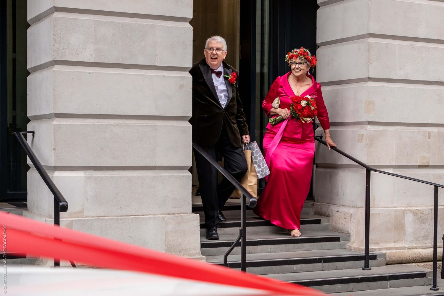Two individuals in formal attire descend stone steps at an elegant building entrance with a red carpet.