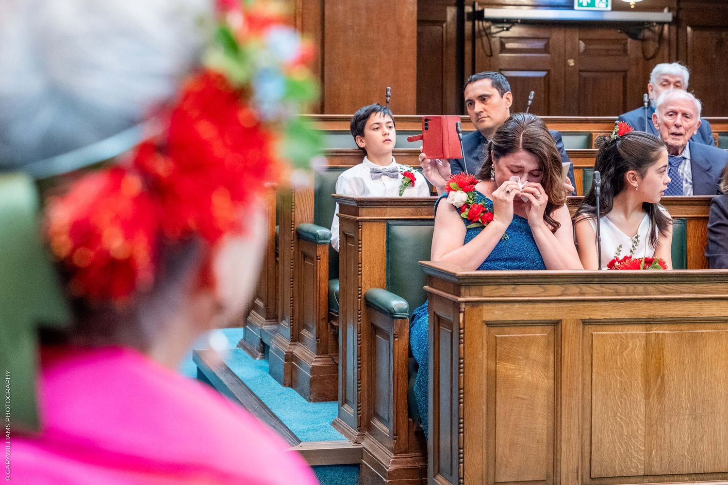 Wedding guests seated in wooden church pews watch attentively during ceremony as someone in bright pink stands nearby.