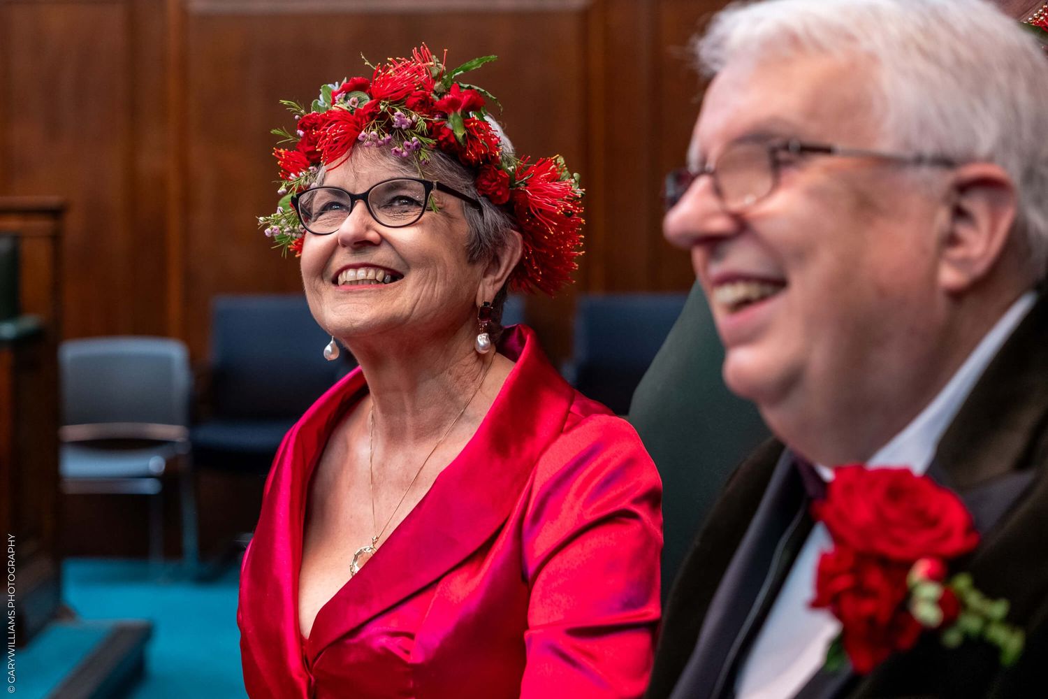 A couple wearing red attire and red floral decorations smile together during a celebratory event.