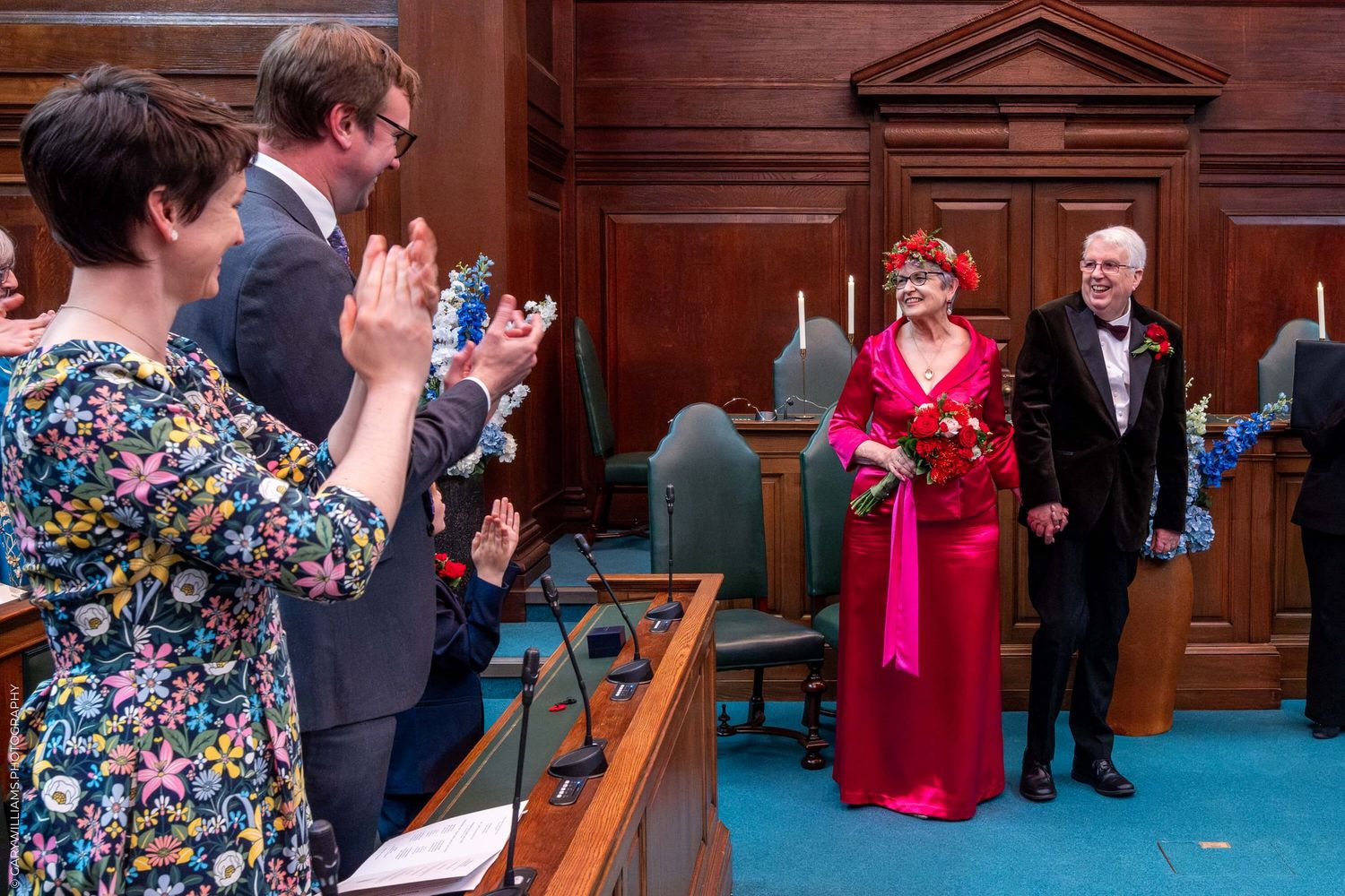 People applaud during a formal event in a traditional legislative chamber with wooden paneling and blue carpeting.
