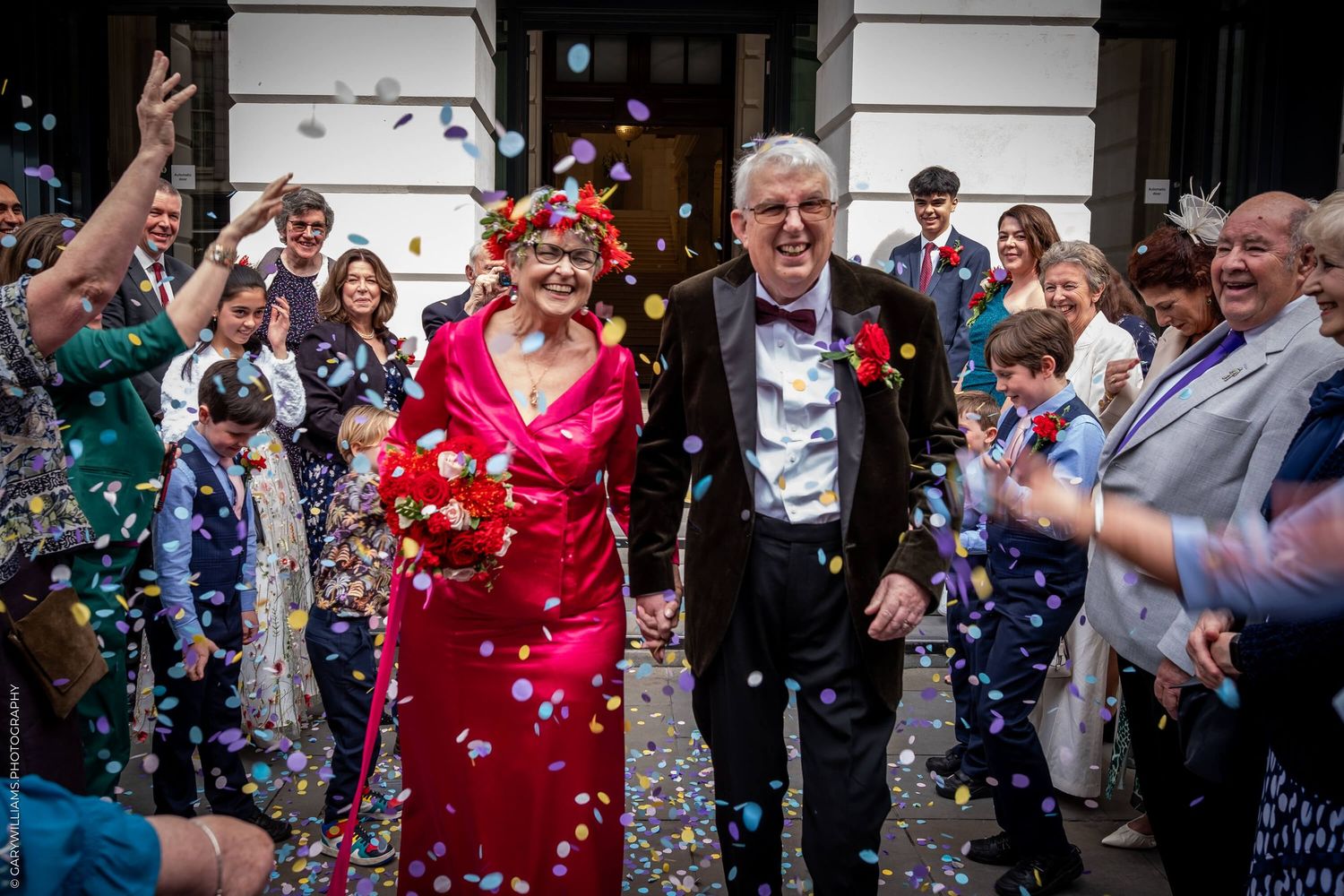 Wedding guests throw colorful confetti at a couple exiting the venue in their formal red and black attire.