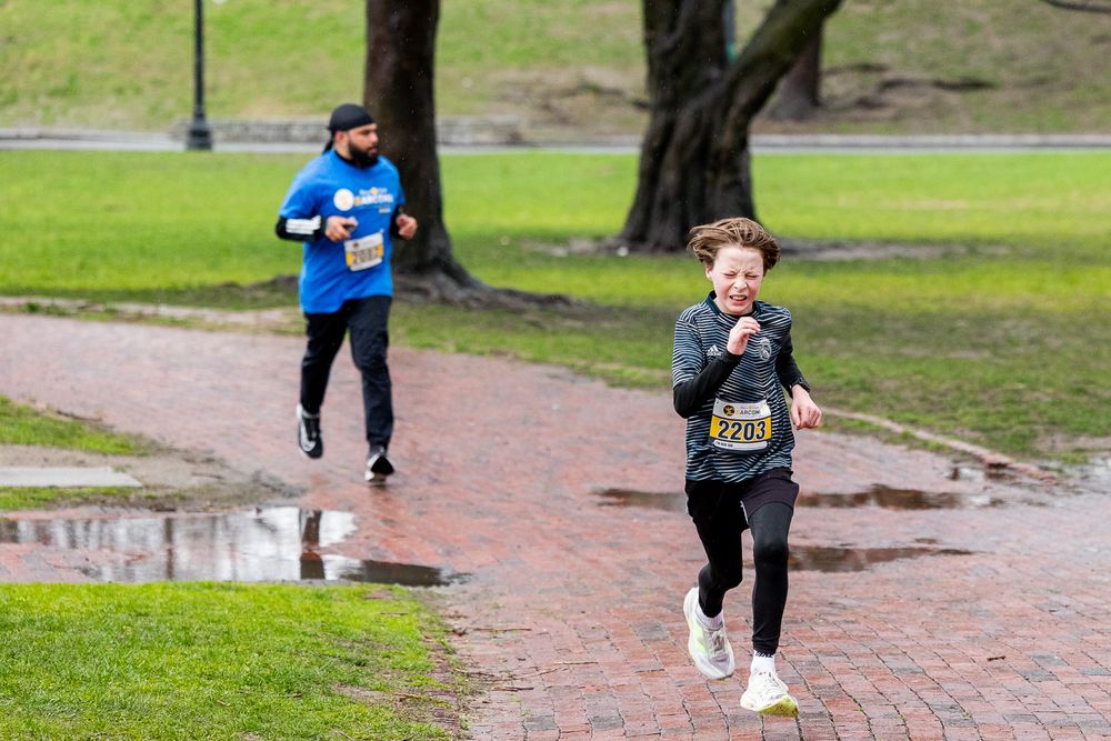 Two runners jog along a wet brick path in a park during what appears to be a race event.
