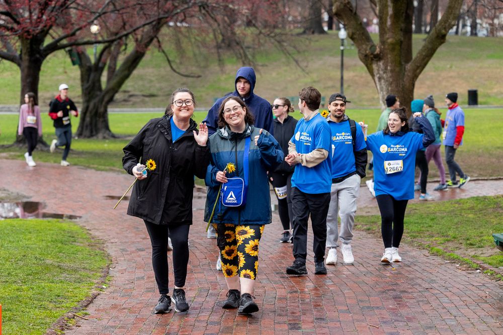 Group of runners in blue shirts and athletic gear jogging along a brick path through a park on an overcast day.