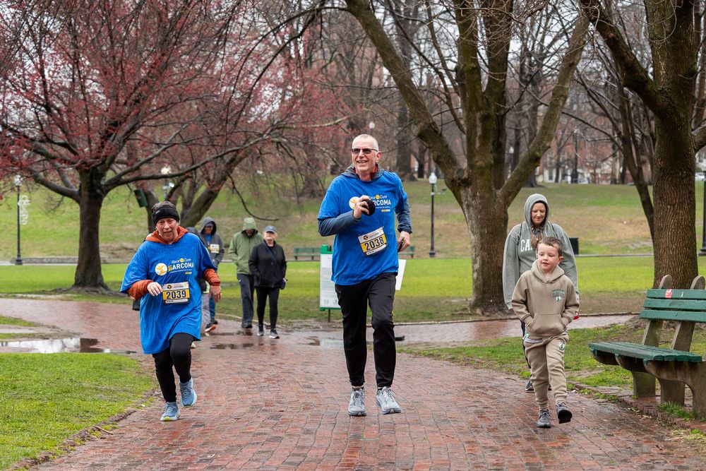 Runners in blue shirts jog along a brick path in a park with blooming cherry trees on a spring day.