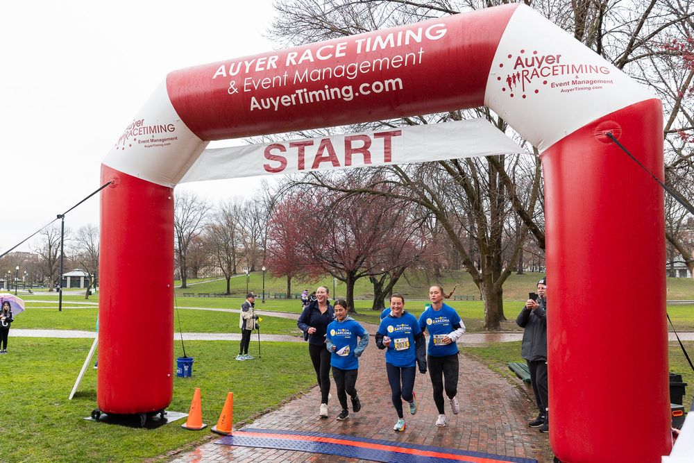 Runners pass through a red inflatable START arch during an outdoor race event in a park setting.