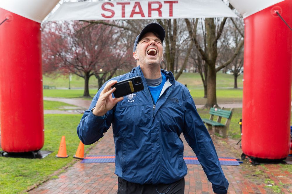 Participant gestures while crossing start line at outdoor running event with red inflatable arch.