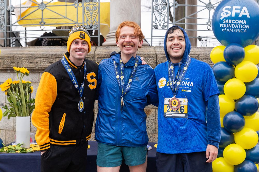 Three runners pose together at a race finish line with balloons and medals near an SFA branded backdrop.