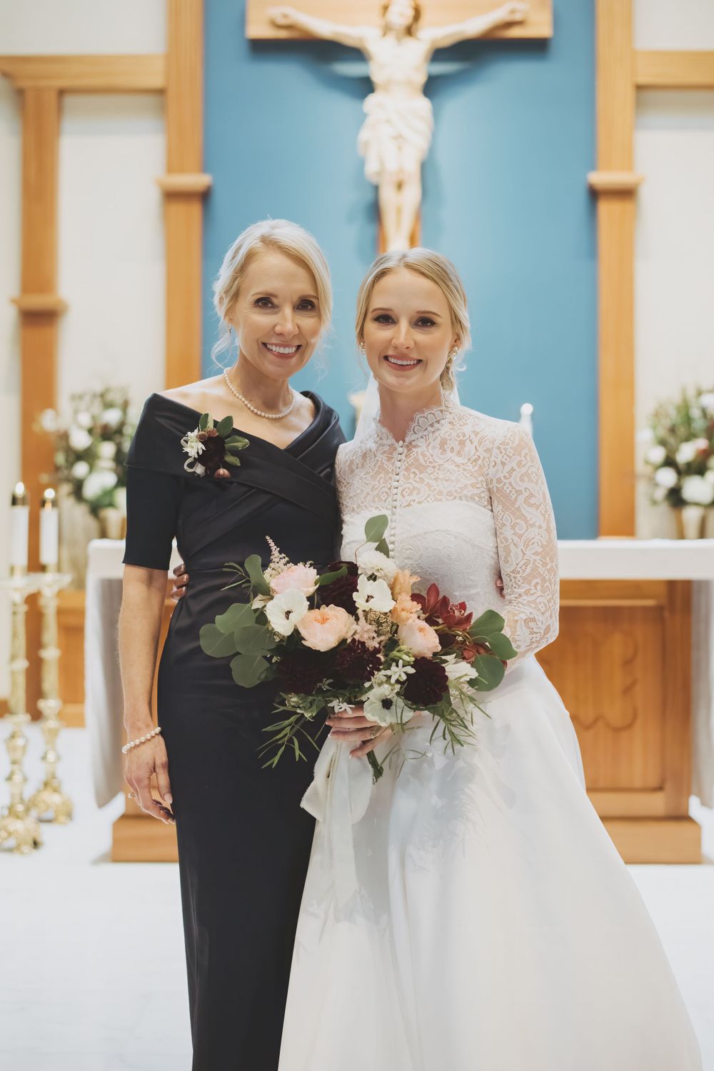 A bride and her mother pose in a church with a crucifix and altar in the background.