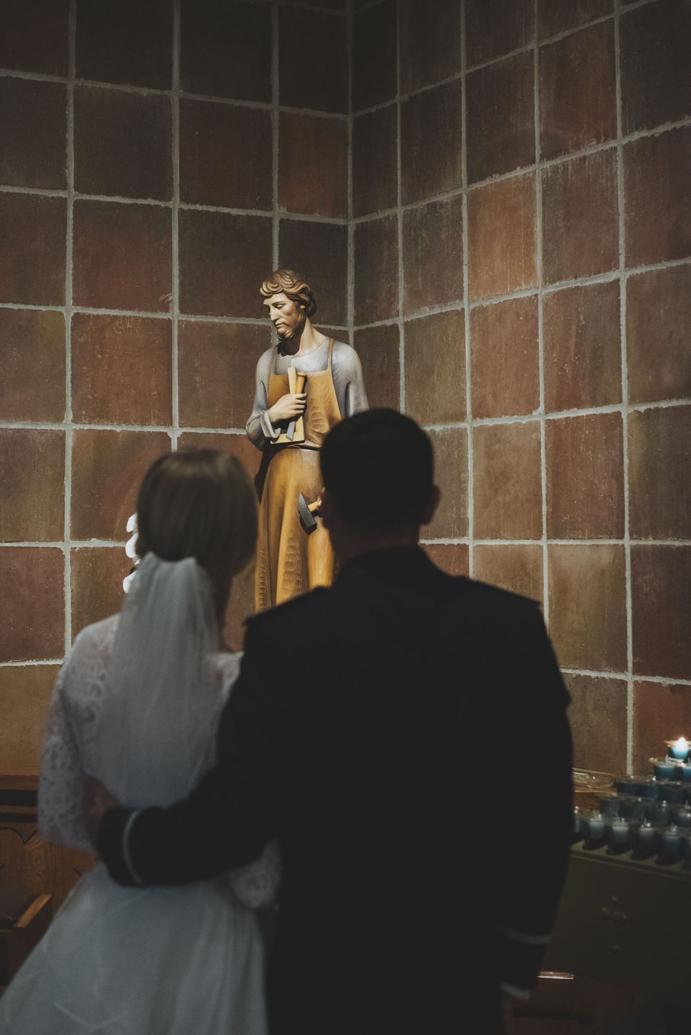 Couple stands before a statue of Saint Joseph the Worker in a church corner during what appears to be a wedding ceremony.