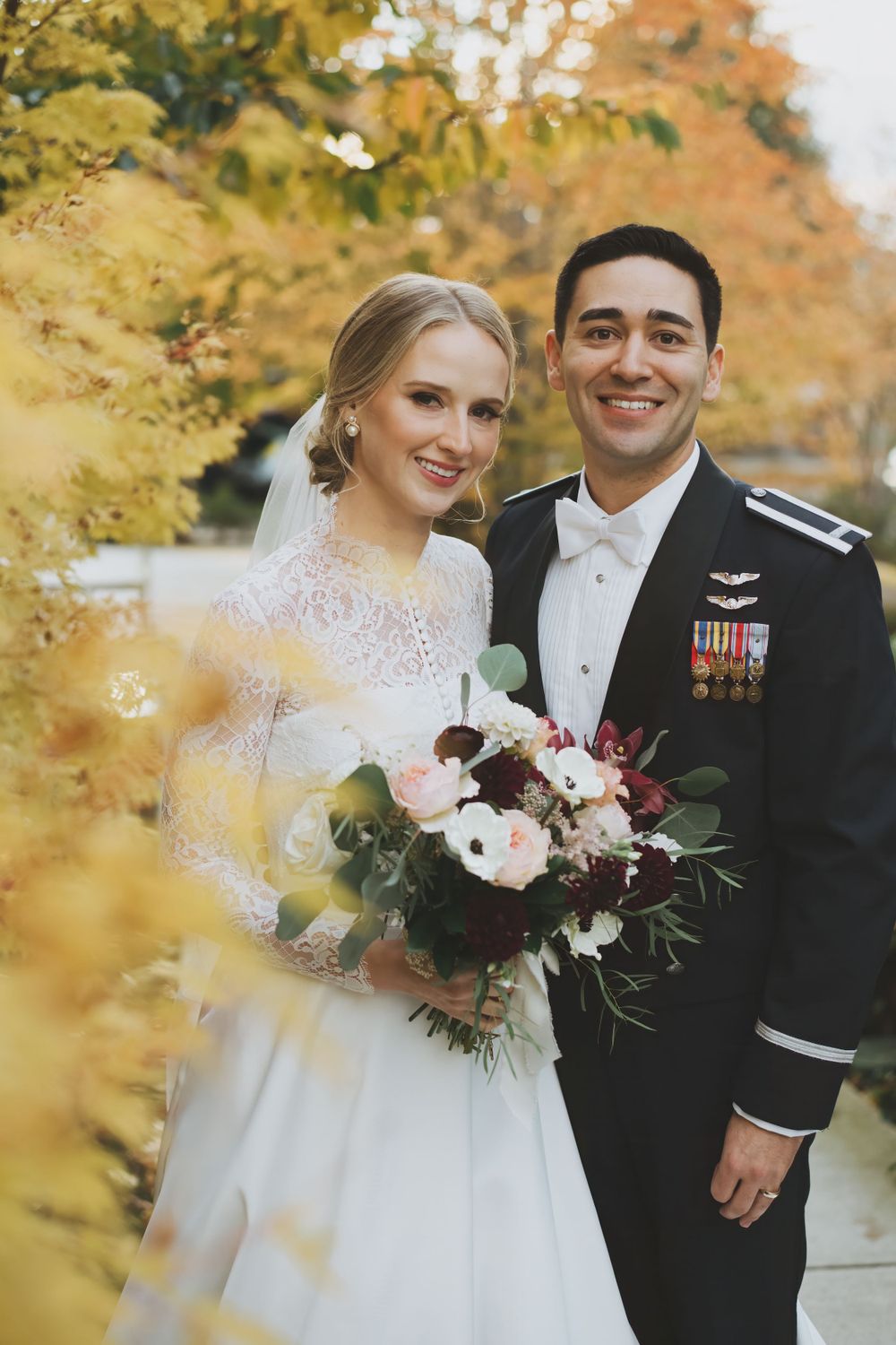 A newlywed couple stands among fall foliage with the bride holding a burgundy and pink bouquet.