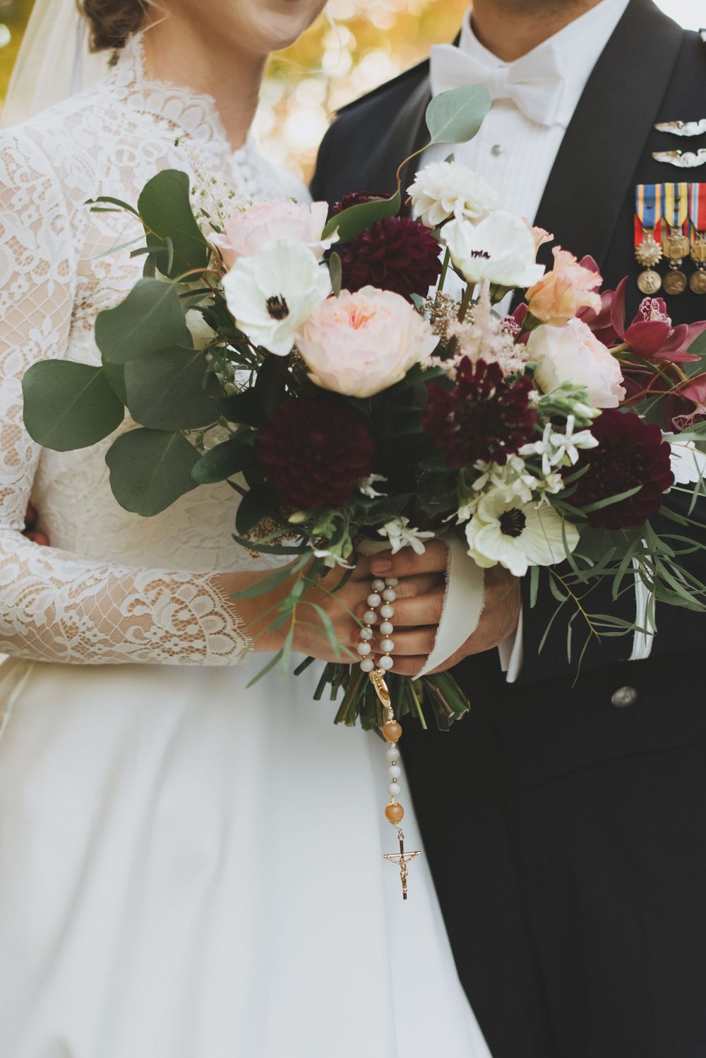 Close-up of wedding bouquet with burgundy and white flowers and rosary held between bride in lace dress and groom in military uniform.
