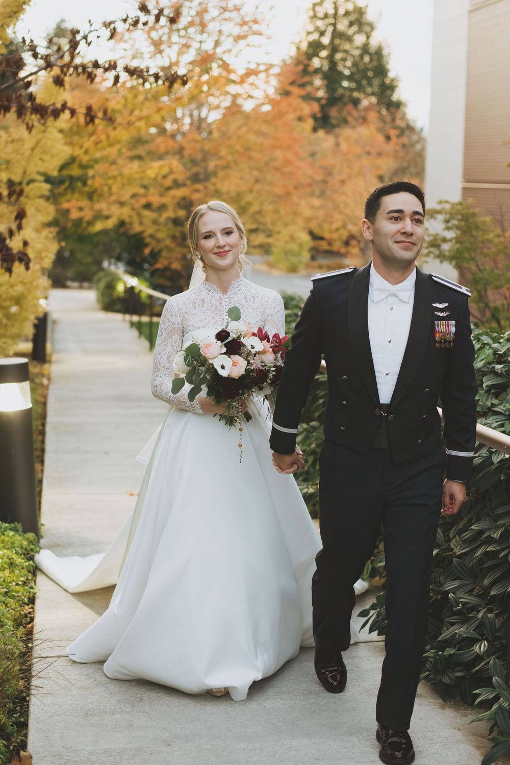 A couple in wedding attire takes a stroll along an autumn path surrounded by trees with yellow foliage.