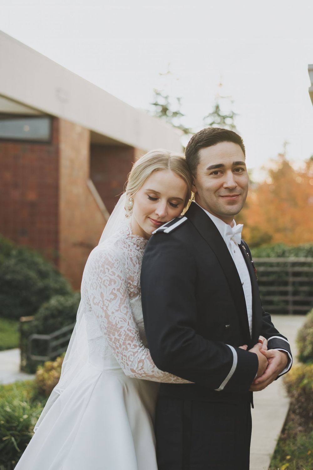 A bride and groom pose together outdoors during an autumn wedding with warm lighting at sunset.
