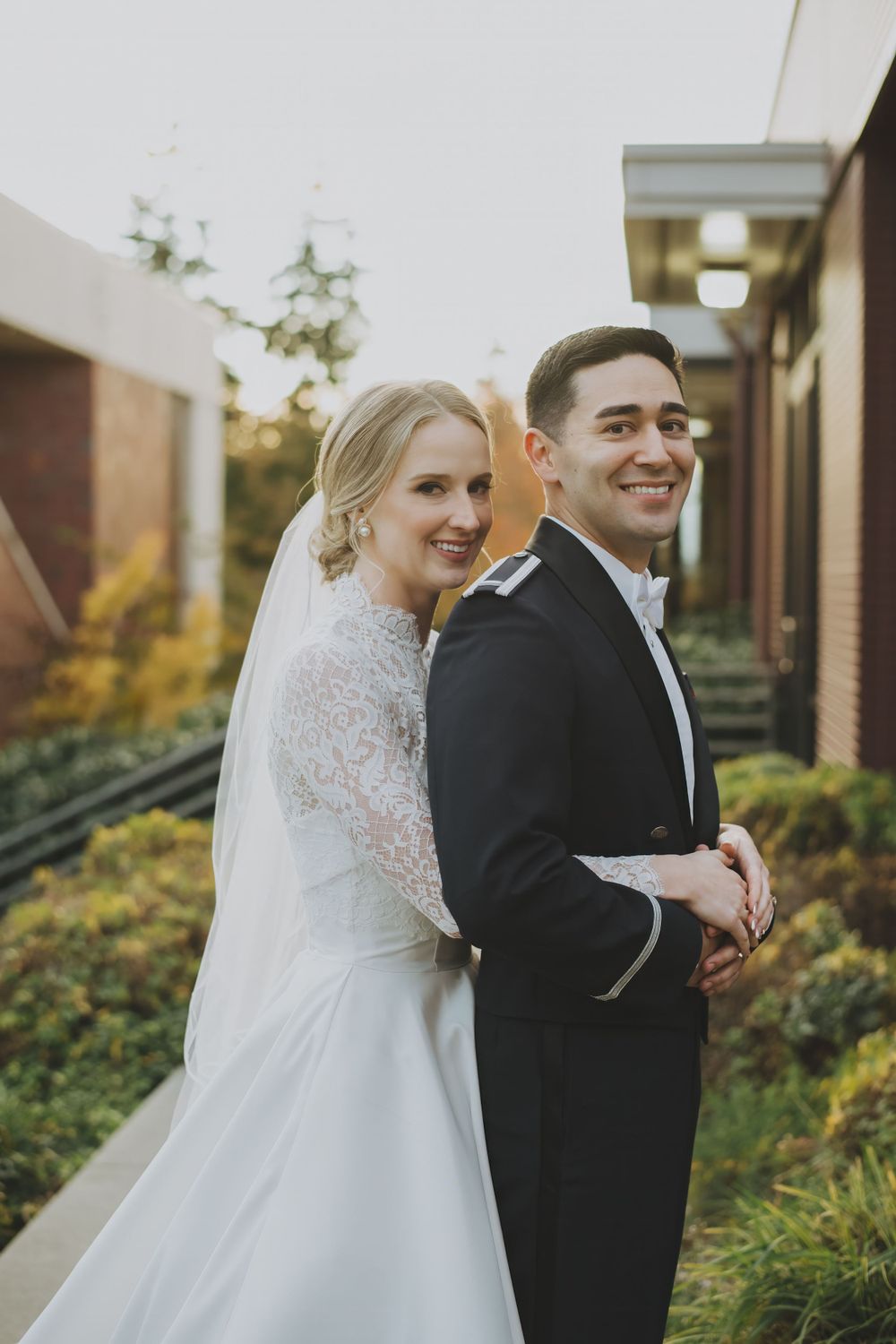 A couple in wedding attire embrace and smile in a garden setting during golden hour.