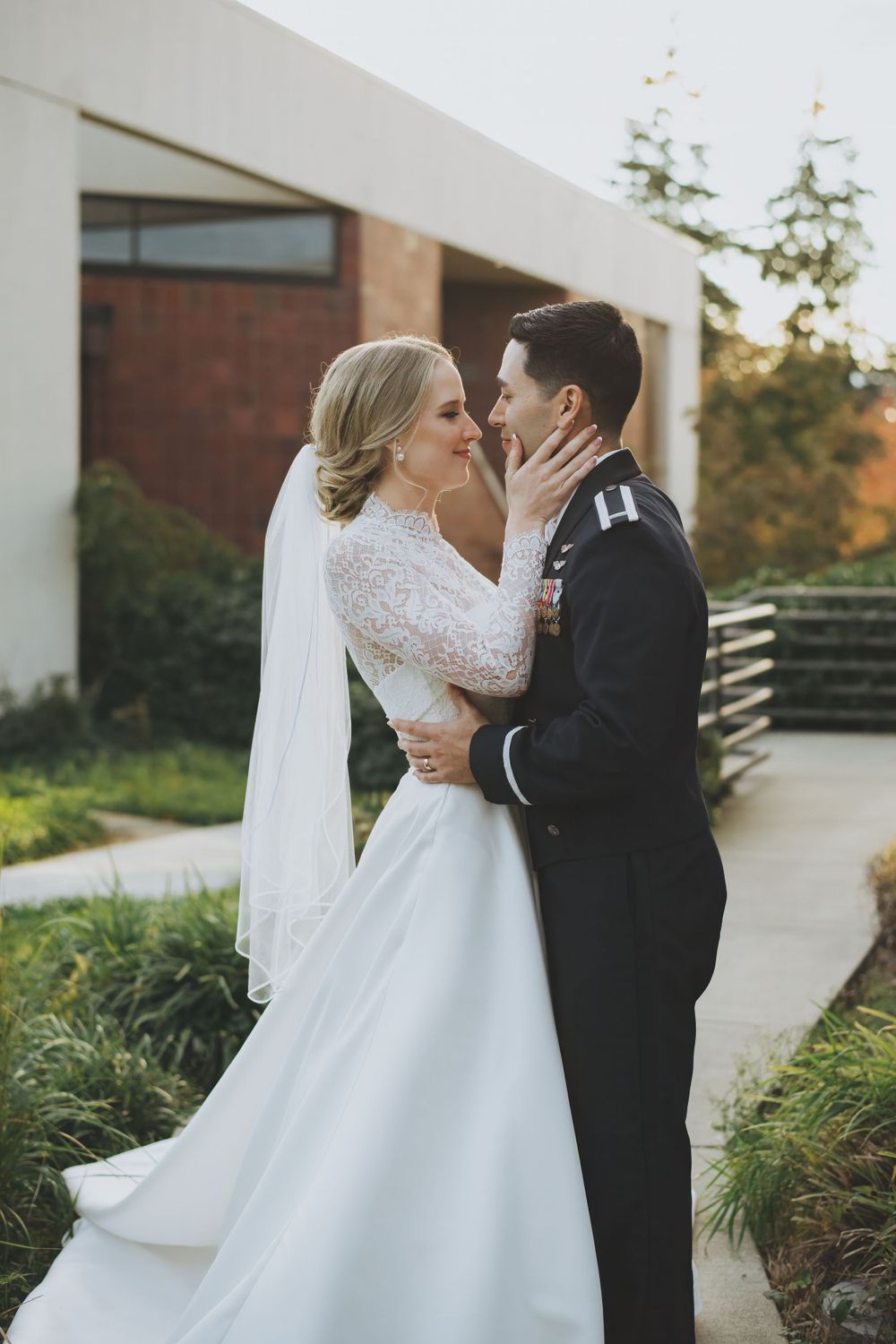A romantic wedding couple embraces outdoors during sunset at a modern venue with greenery in the background.
