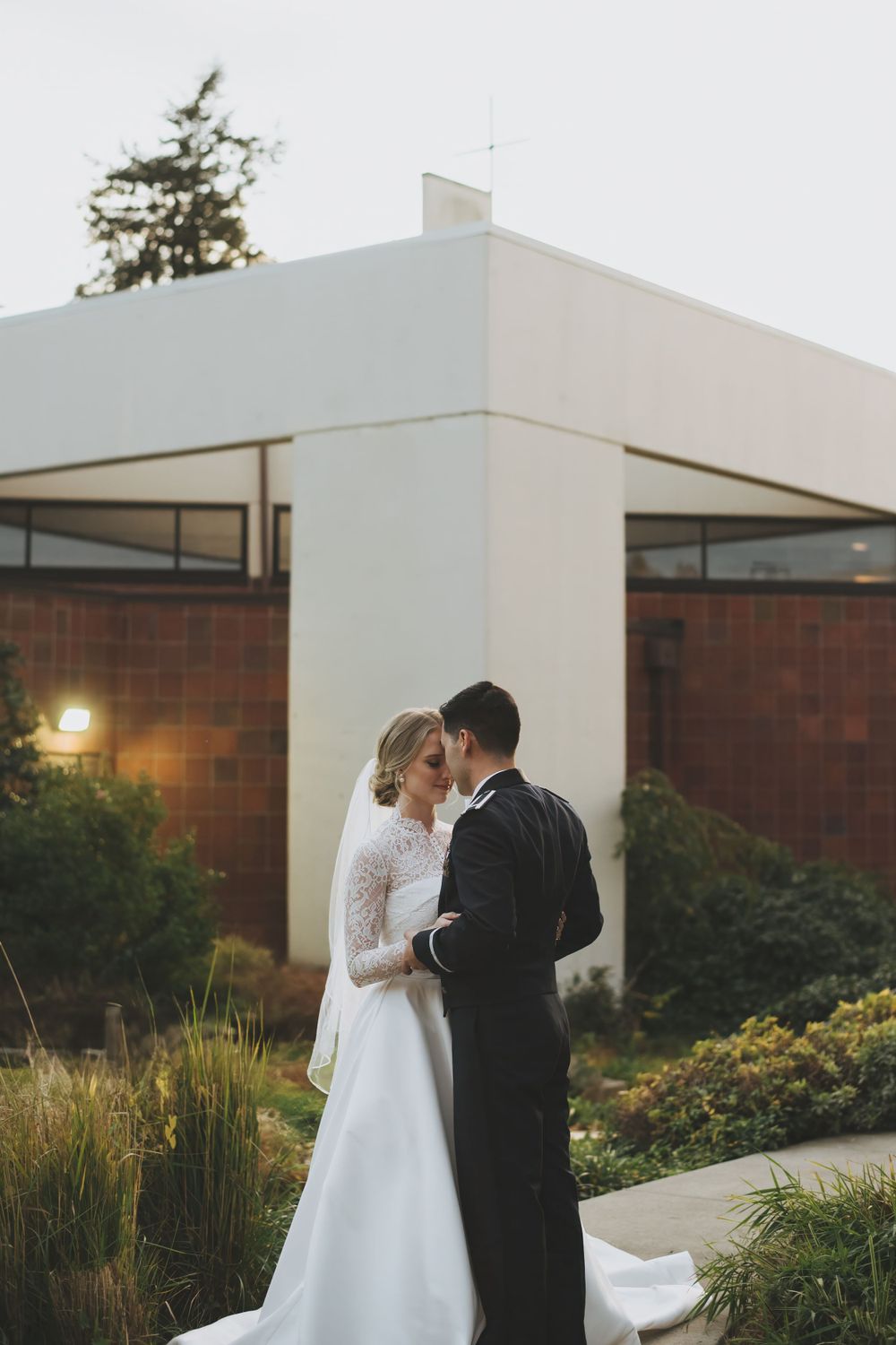 A romantic wedding couple embraces outside a modern building at sunset, surrounded by ornamental grasses.
