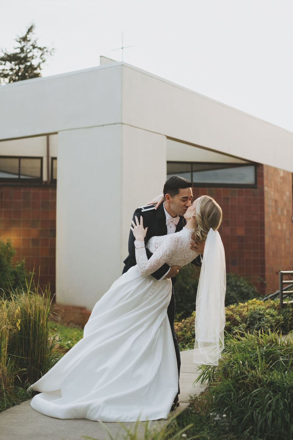 A romantic wedding photo showing a couple in formal attire sharing a kiss outside a modern building with landscaping.