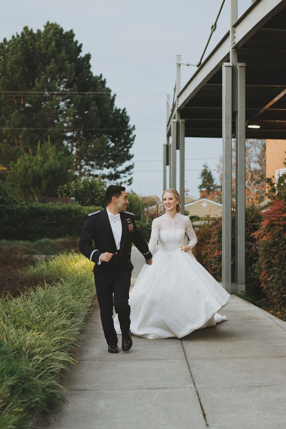 A couple in formal wedding attire running together on a sidewalk path lined with ornamental grasses.