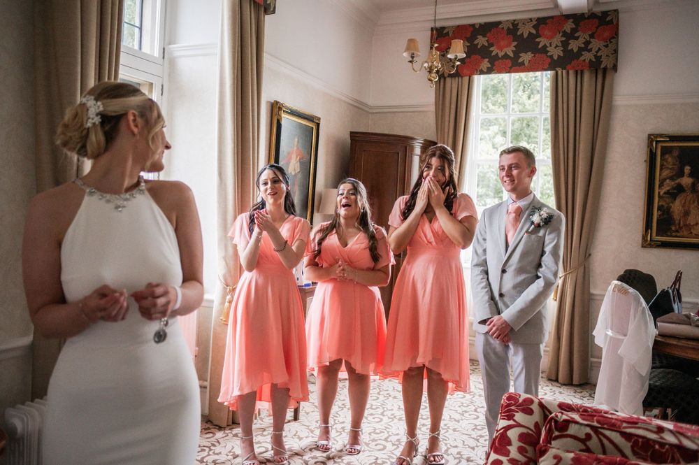 Wedding party in coral dresses react with joy during pre-ceremony preparations.
