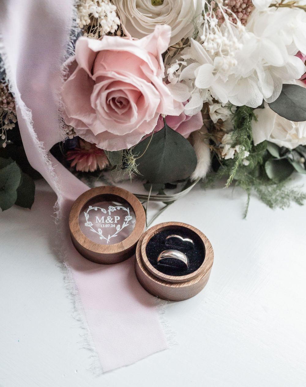 Wedding rings in wooden boxes surrounded by pink and white roses and greenery.