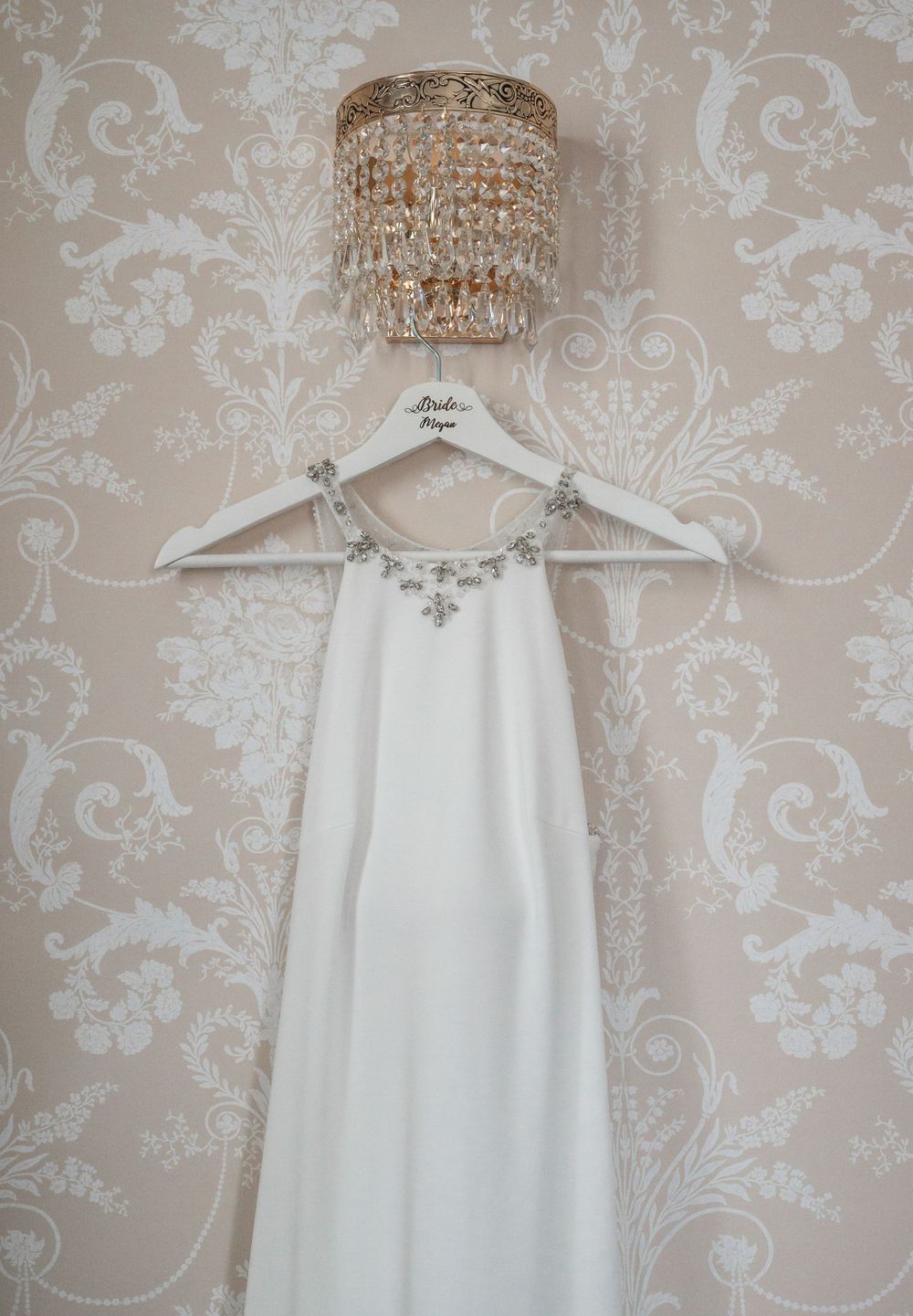 White wedding dress hanging under crystal chandelier against damask wallpaper.