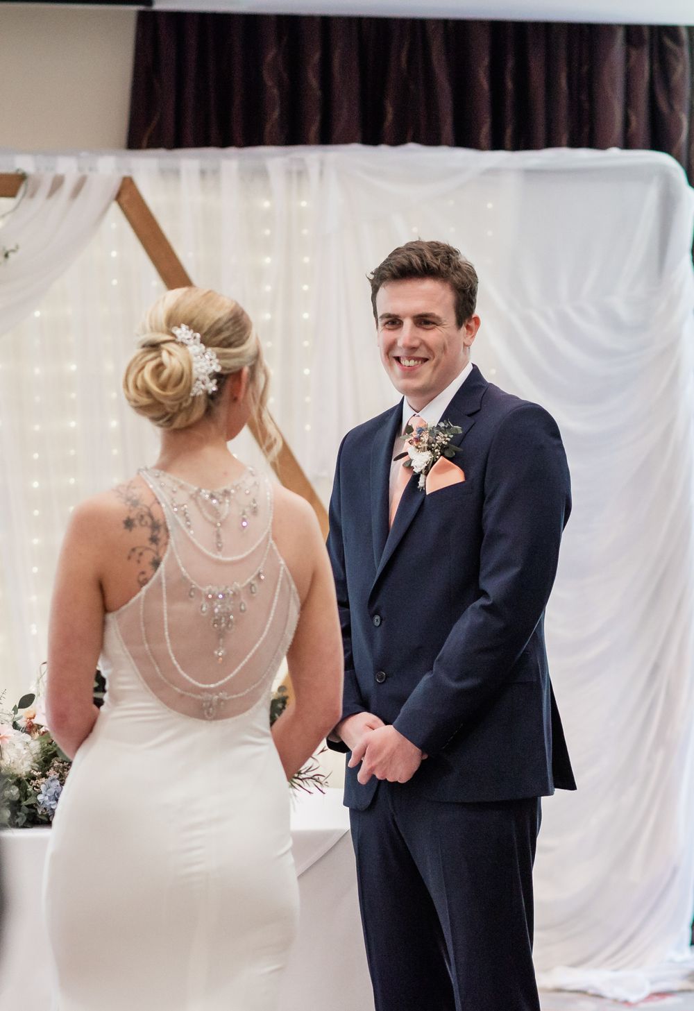 A bride in a fitted white dress with sheer back detail shares a moment with her groom during the ceremony.