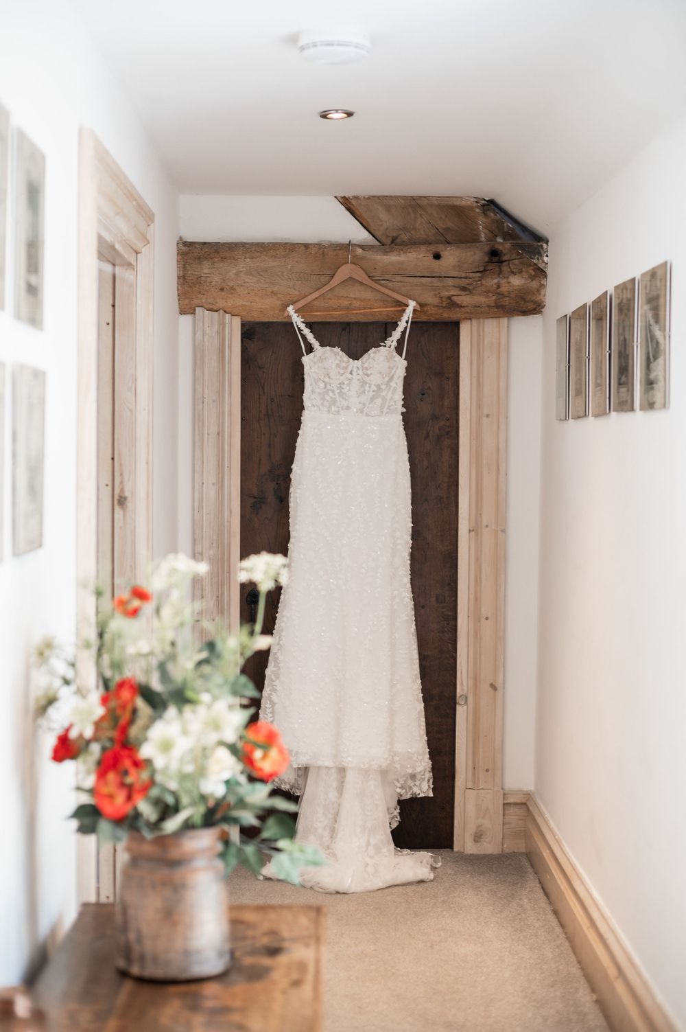 White lace wedding dress hanging in a rustic hallway with floral decorations.