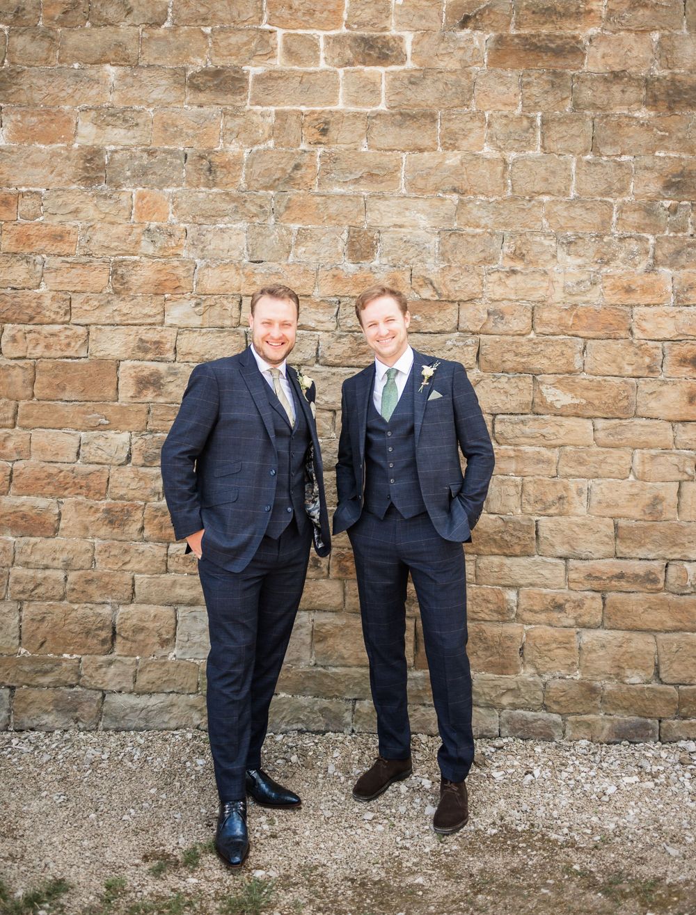 Two men in formal suits share a lighthearted moment against a stone wall background.