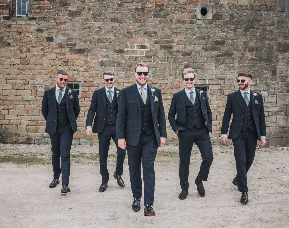 Groomsmen in dark suits and sunglasses walking confidently against stone wall background.