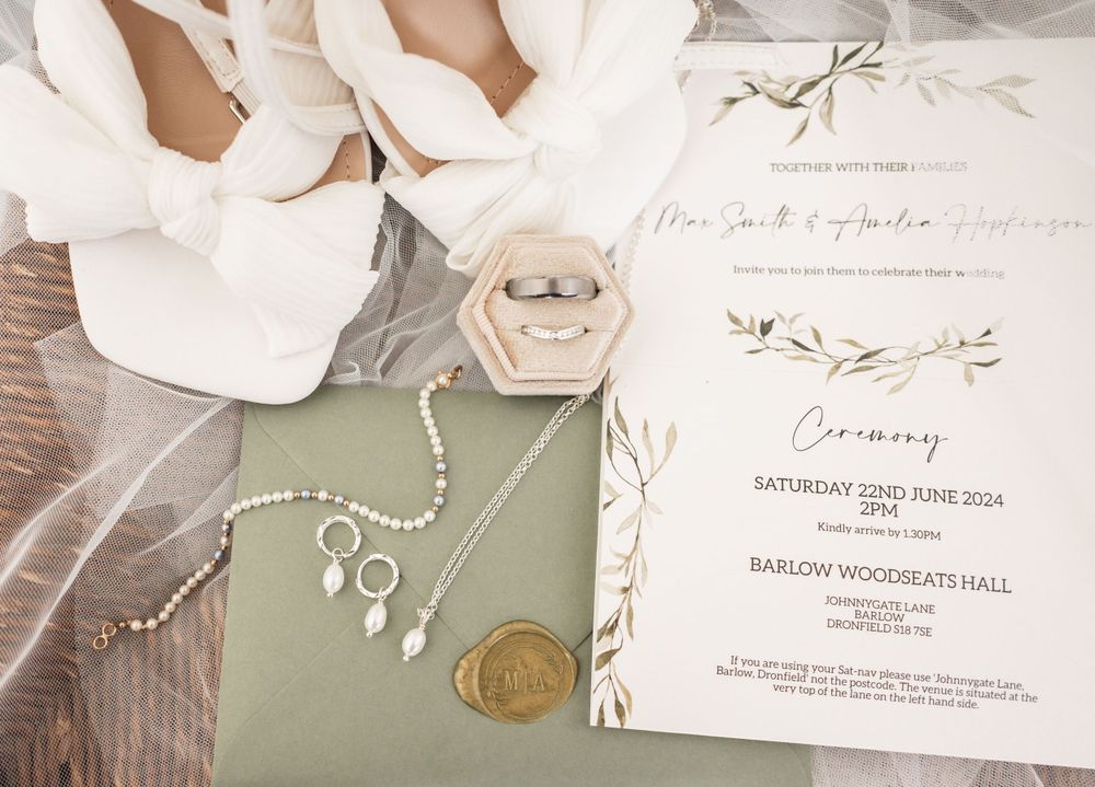 Wedding invitation and jewelry details with white floral elements and sage green accents on a wooden surface.