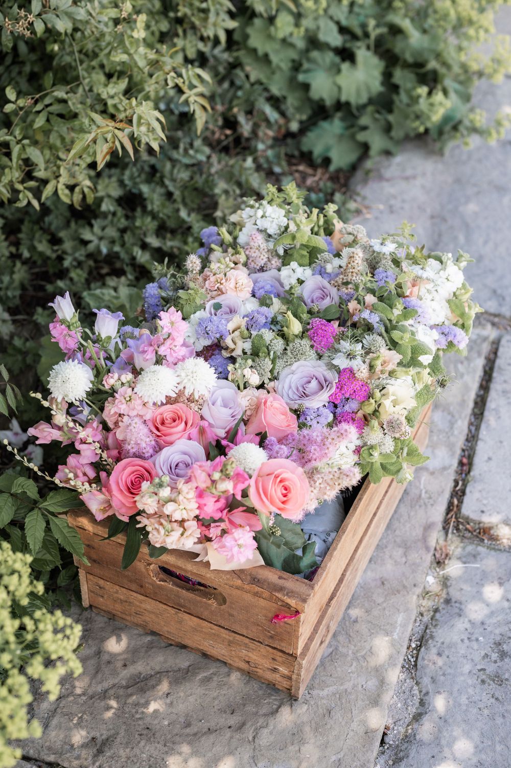 Rustic wooden crate filled with pink, purple, and white flowers arranged in a garden setting.