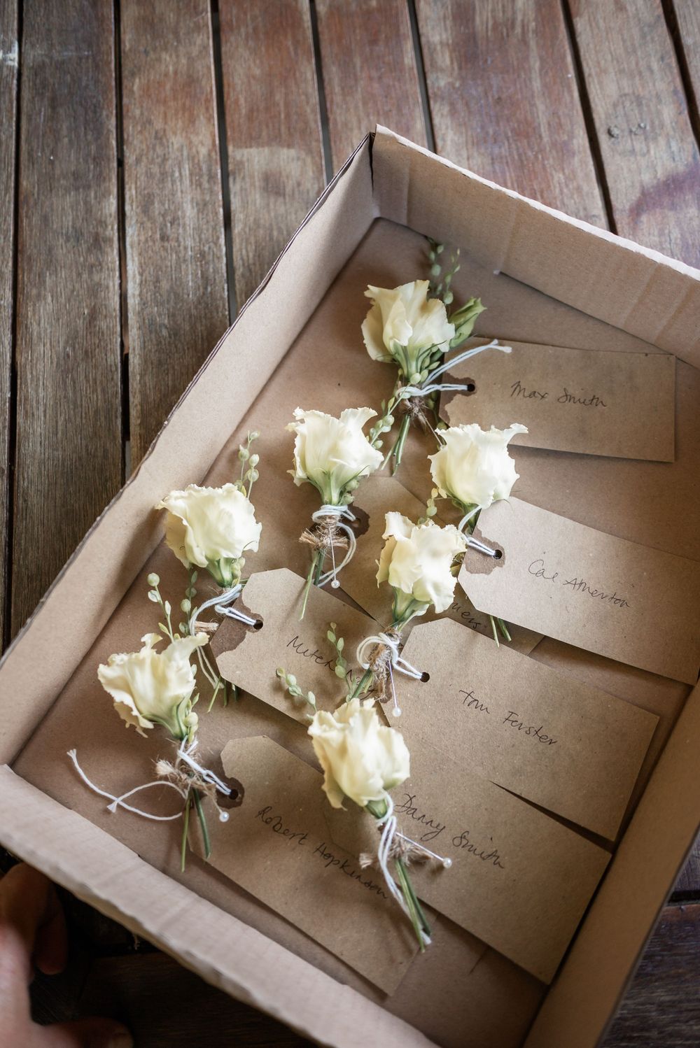 White rose boutonnieres with name cards arranged in a kraft paper box.