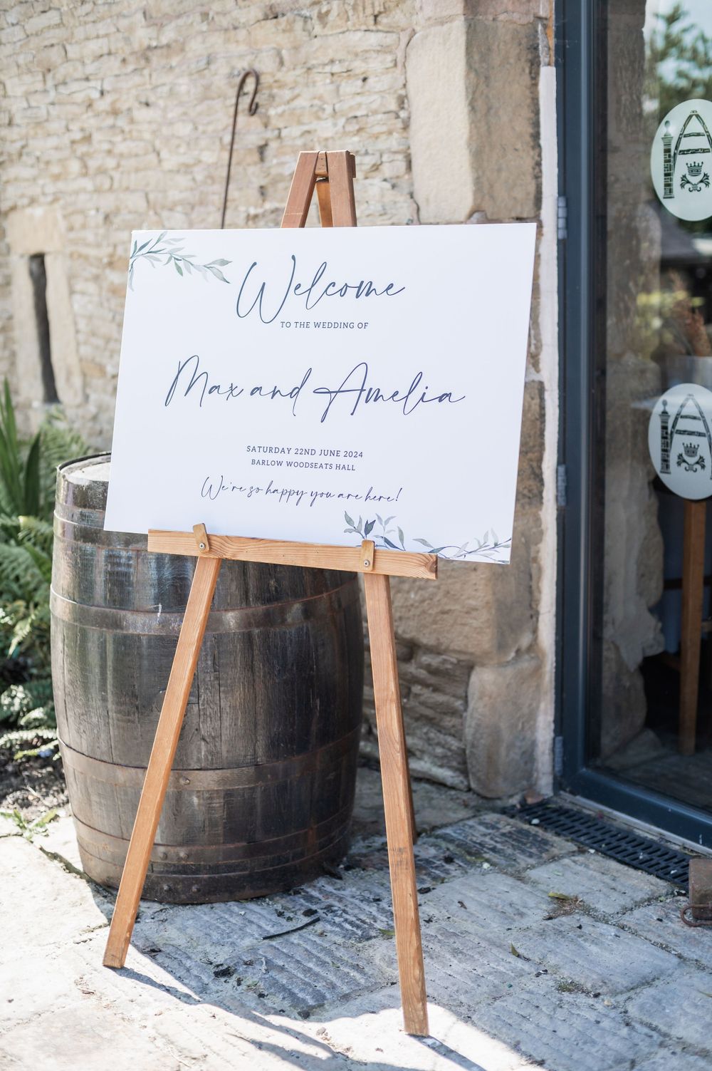 Welcome sign displayed on wooden easel beside vintage barrel at venue entrance.