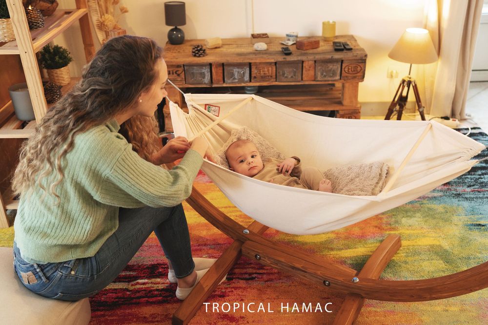A peaceful scene with a wooden baby hammock and caregiver in a cozy living room with warm lighting.