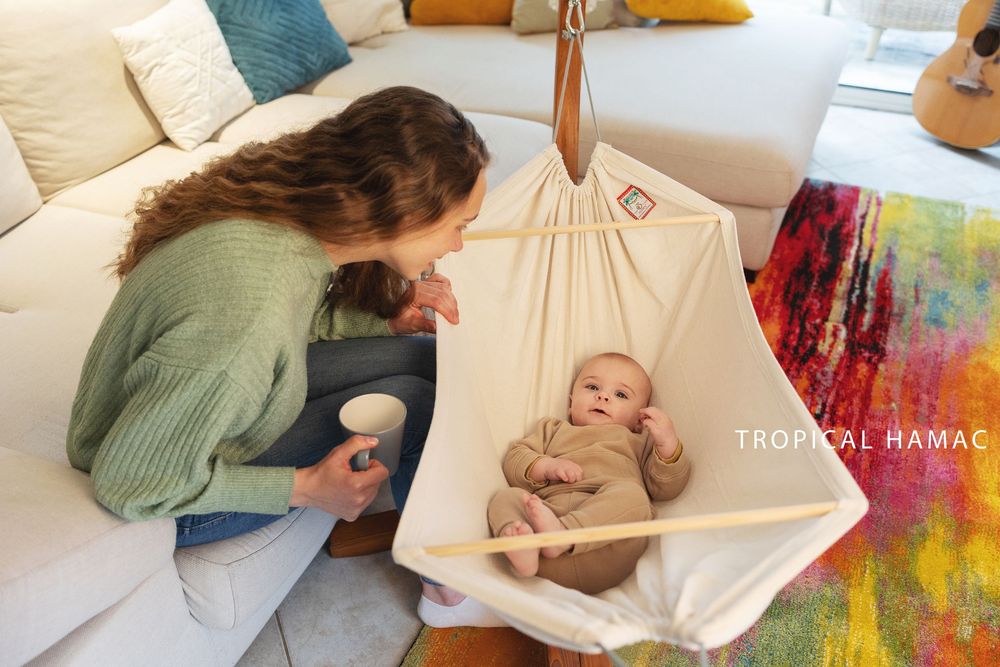 Adult in green sweater leans over to interact with baby resting in suspended hammock in bright room.