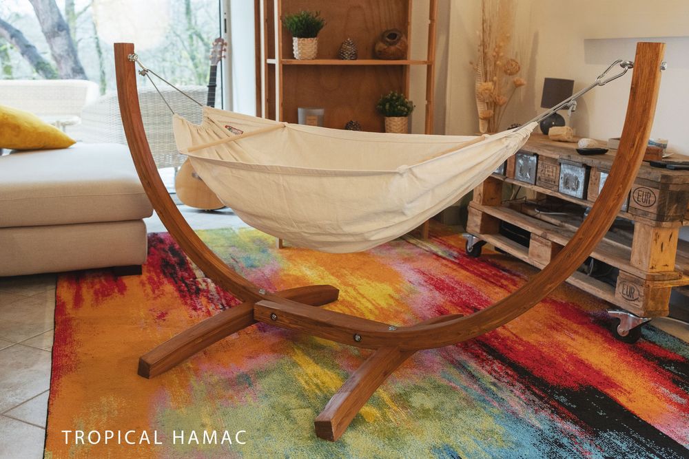 Curved wooden stand hammock with cream fabric displayed on colorful rainbow area rug in modern living room setting.
