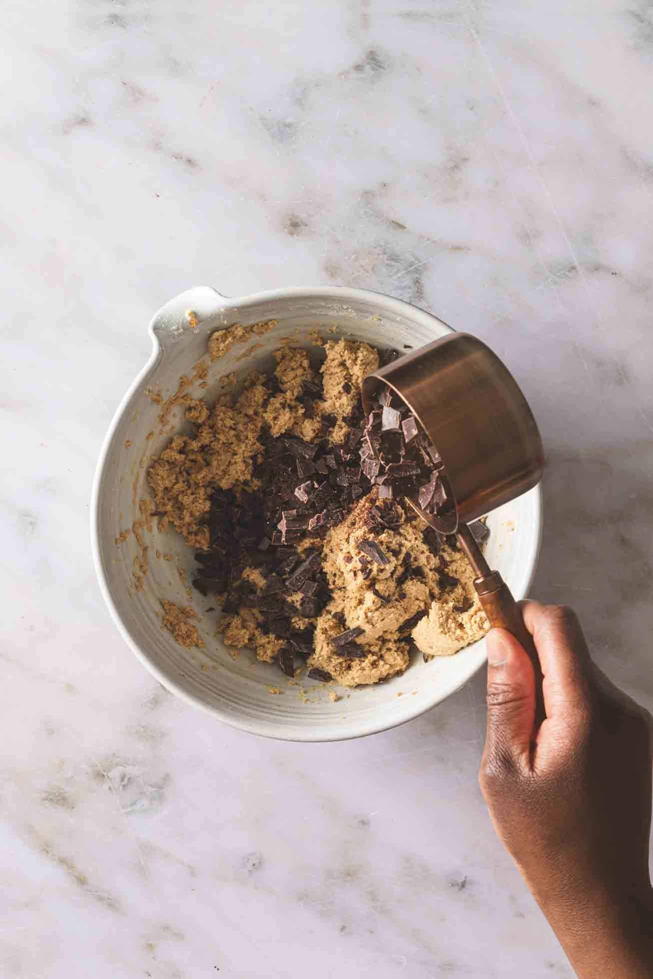 Hand pouring dark chocolate into a bowl of cookie dough mixture on a marble countertop for homemade baking.