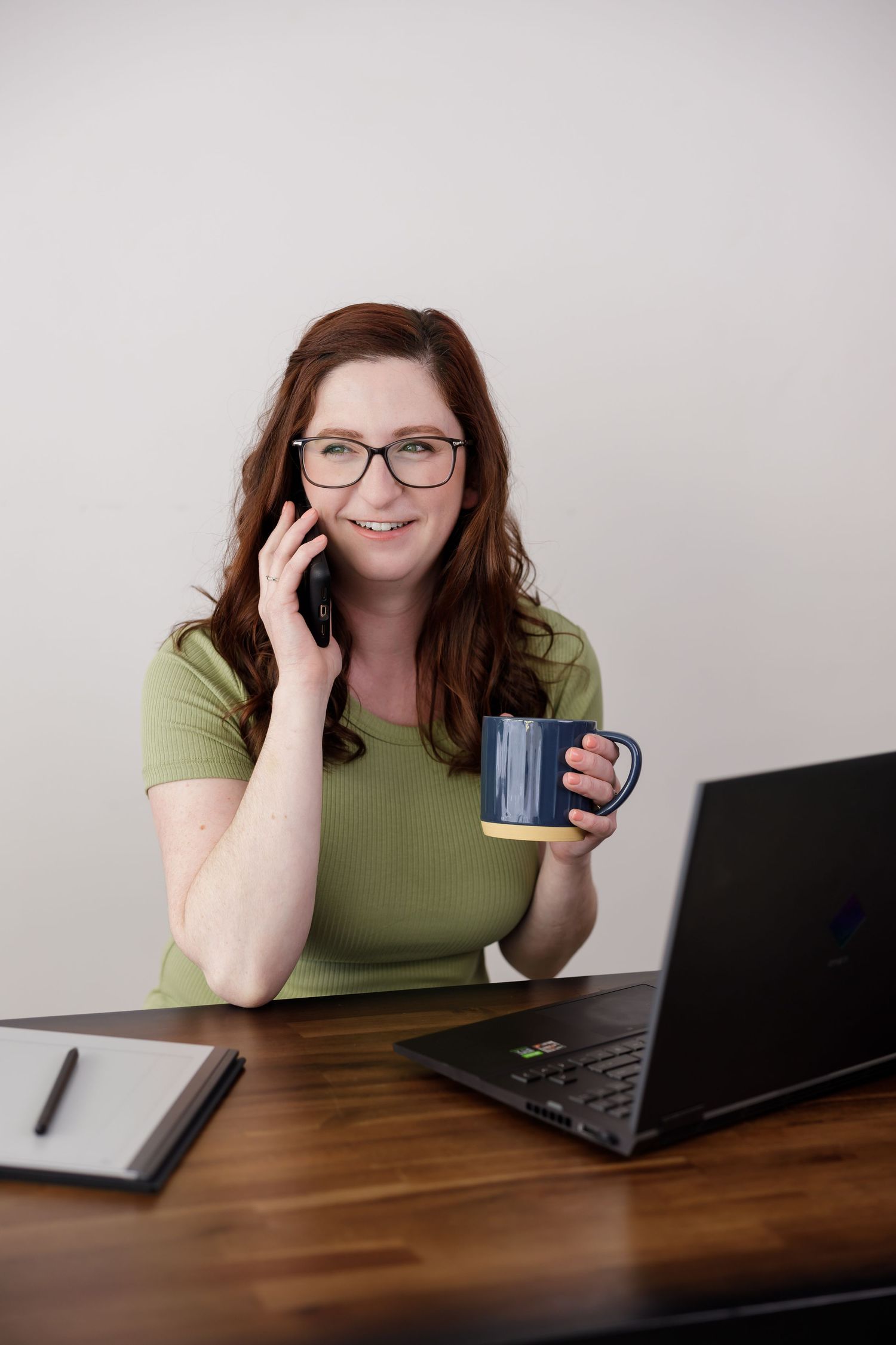 woman graphic designer sitting at desk talking on phone with mug in hand and laptop on desk for lifestyle personal branding in Bothell, WA