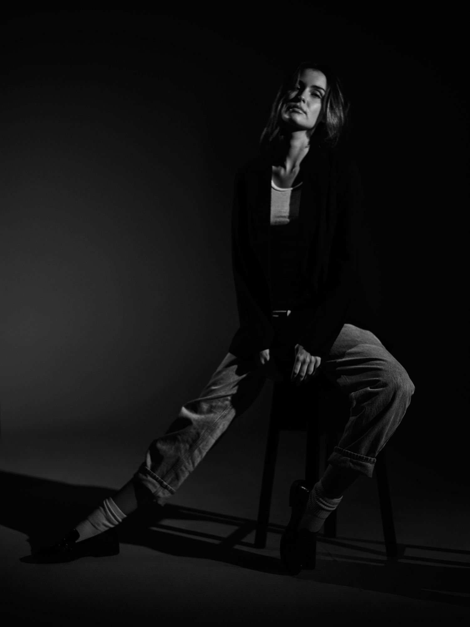 Artistic black and white fashion portrait of a person in dark clothing posing dramatically against a dark background.